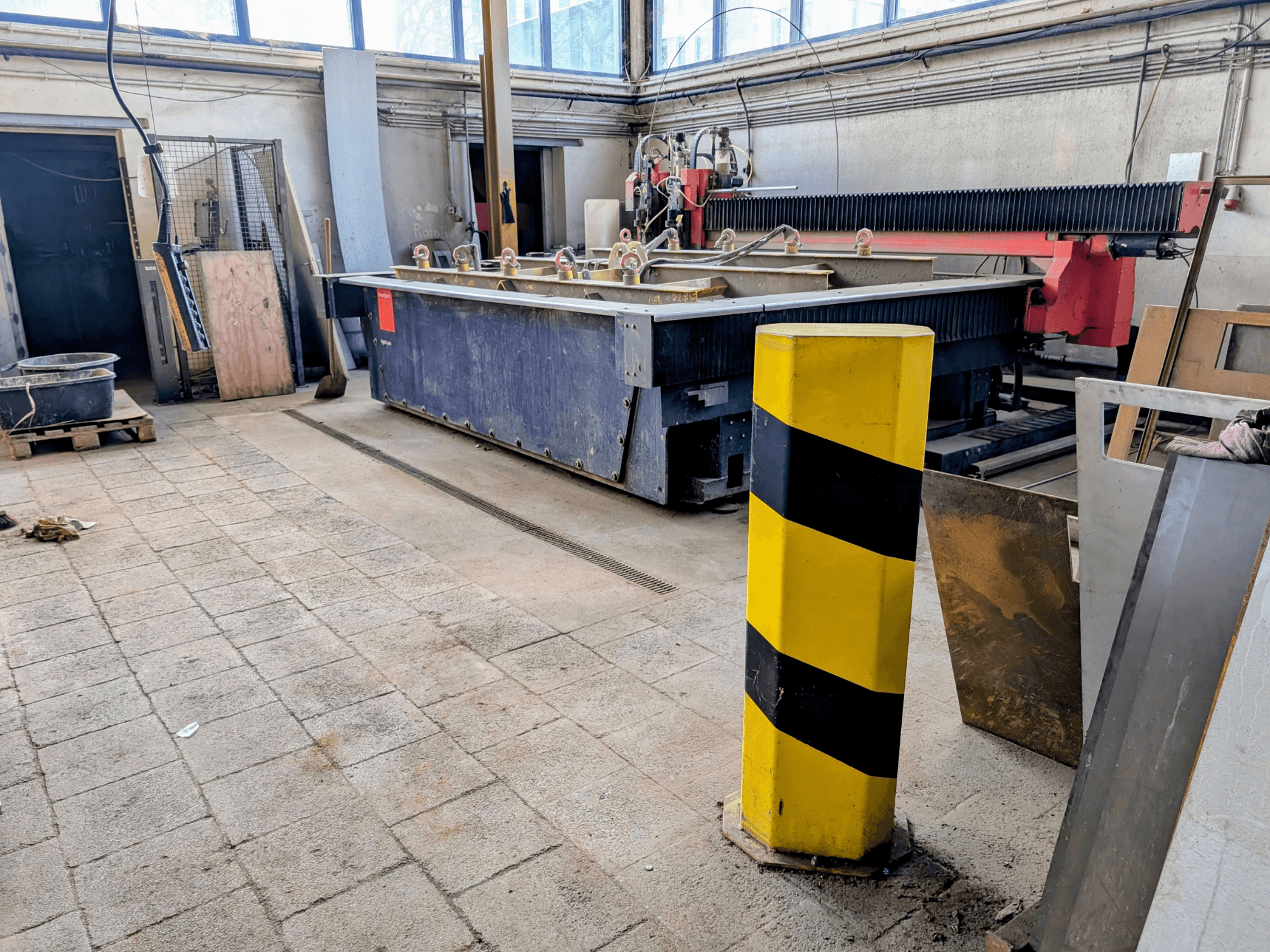 Bystronic Byjet L 2030 -vesisuihkuleikkauskone korjaamolla, sivukuva, keltainen turvapylväs ja ympäröivät materiaalit.