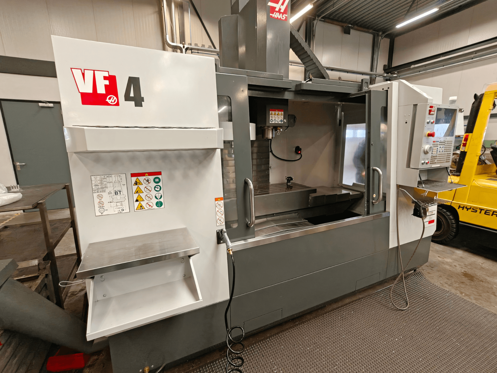 HAAS VF-4 -pystytyöstökeskus, etukuva, jossa on ohjauspaneeli, työalue ja sivussa olevat turvatarrat.