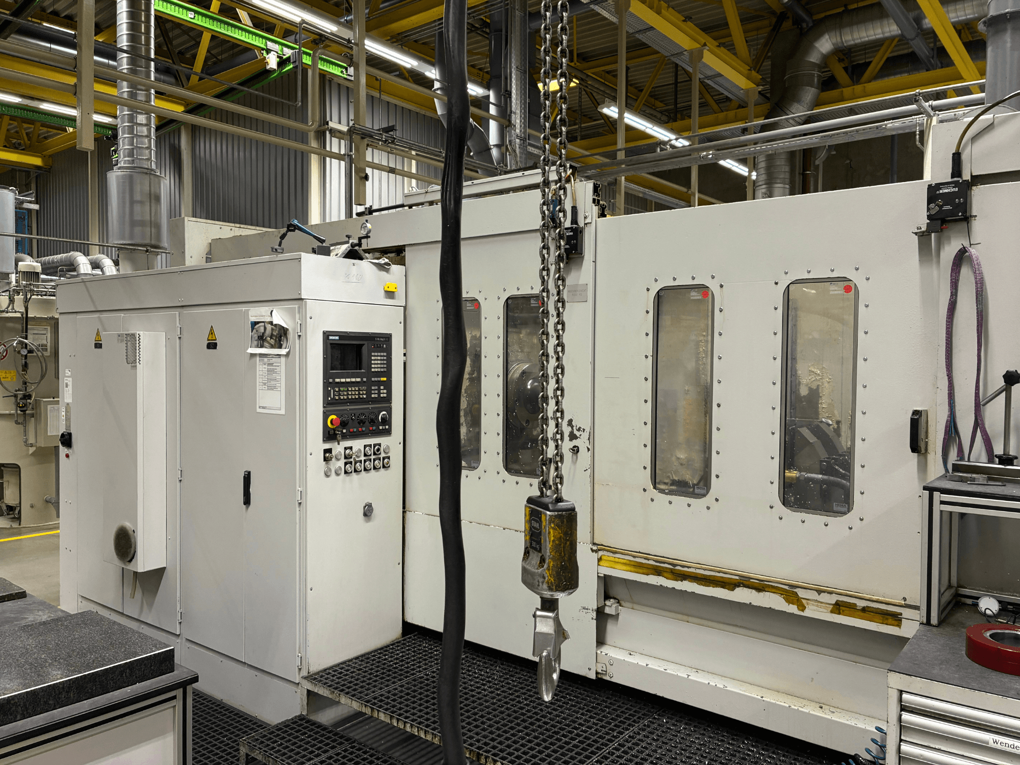 Teollinen CNC-kone ohjauspaneelilla, sivukuva; ikkunoita, ketjuja ja metalliosia tehdasympäristössä.