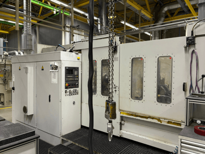 Teollinen CNC-kone ohjauspaneelilla, sivukuva; ikkunoita, ketjuja ja metalliosia tehdasympäristössä.